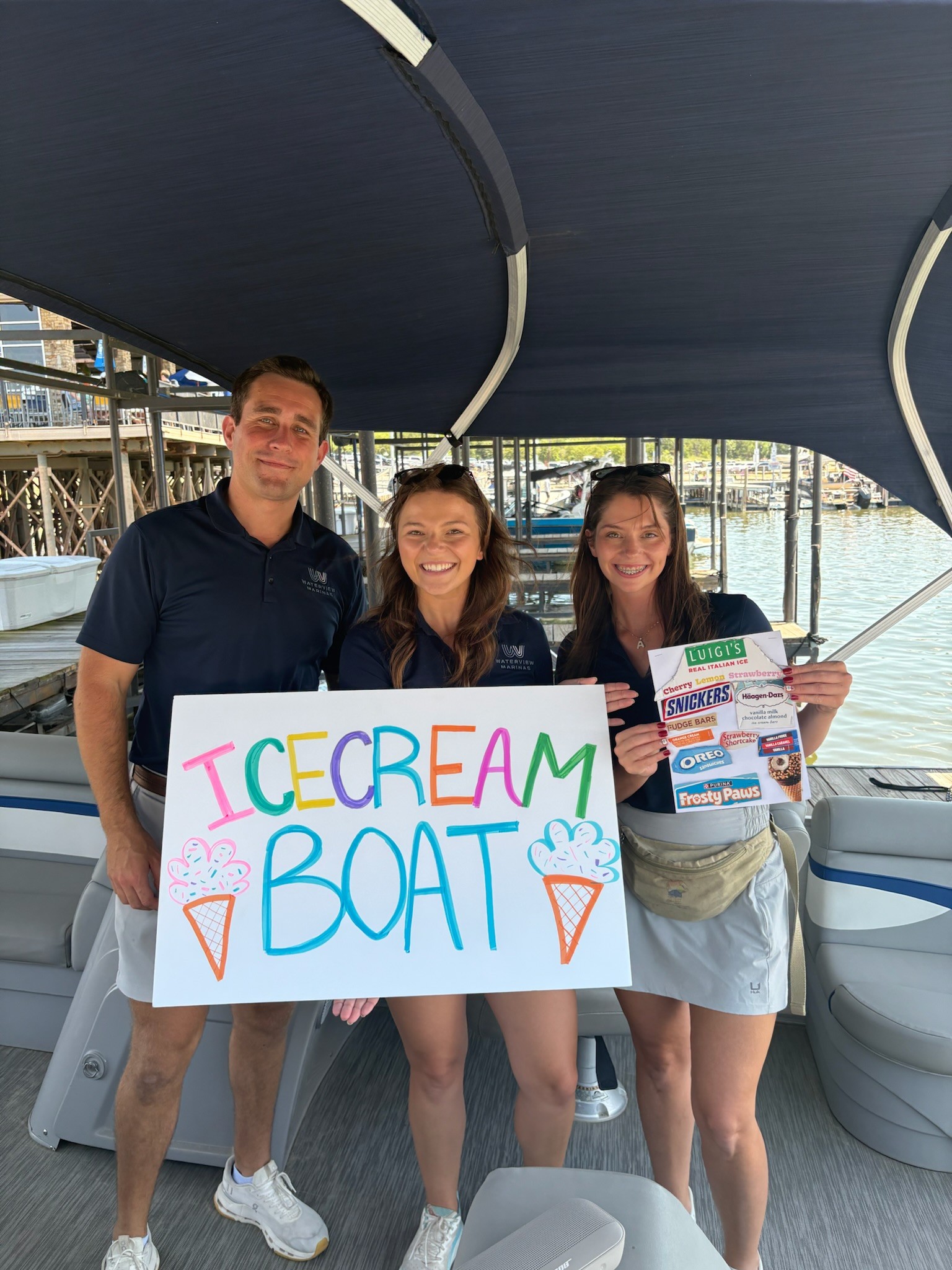 Three people stand on a boat holding a colorful sign that reads "Ice Cream Boat," with blue water and a dock in the background.