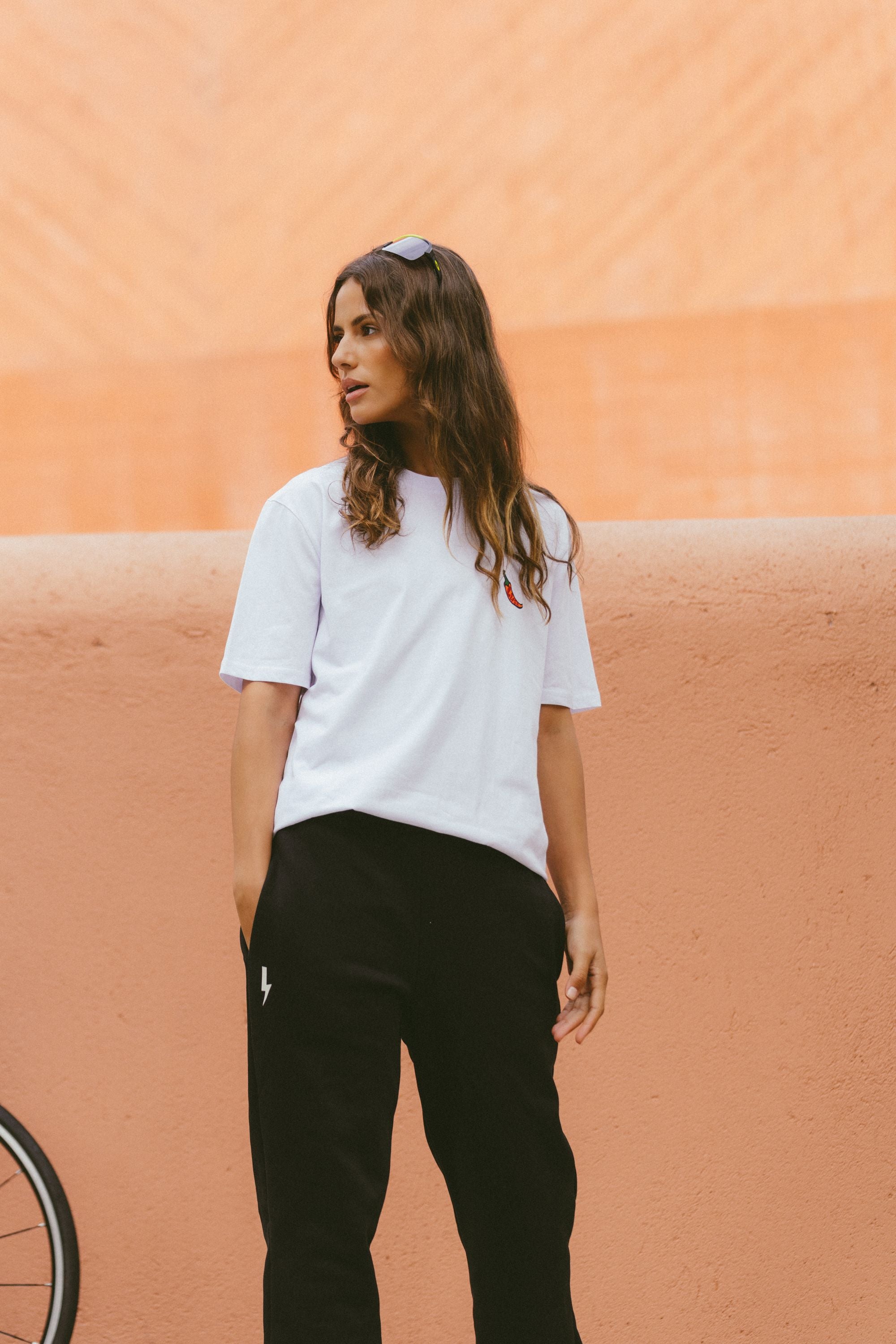 A-woman-with-long-wavy-hair-looking-off-to-the-side-wearing-a- white-t-shirt-with-a-small-chili- pepper-embroidery-on-the-chest- and-black-sweatpants-with-a-white- lightning-bolt-sign-on-the-left-hip She-is-standing-with-her-hands-in- her-pockets-against-a-warm-terracotta-colored-wall