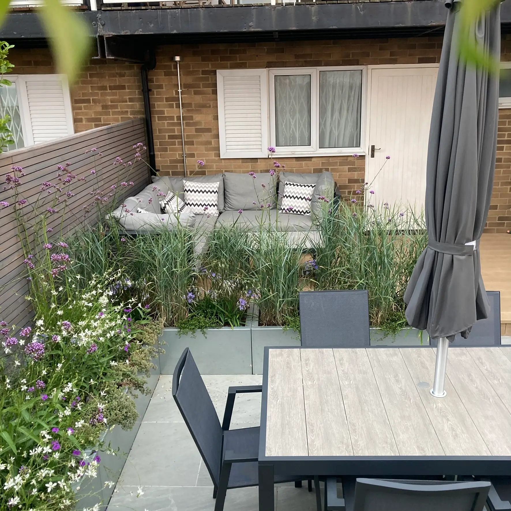 A backyard view featuring a dining table, chairs, and greenery near a house with windows.
