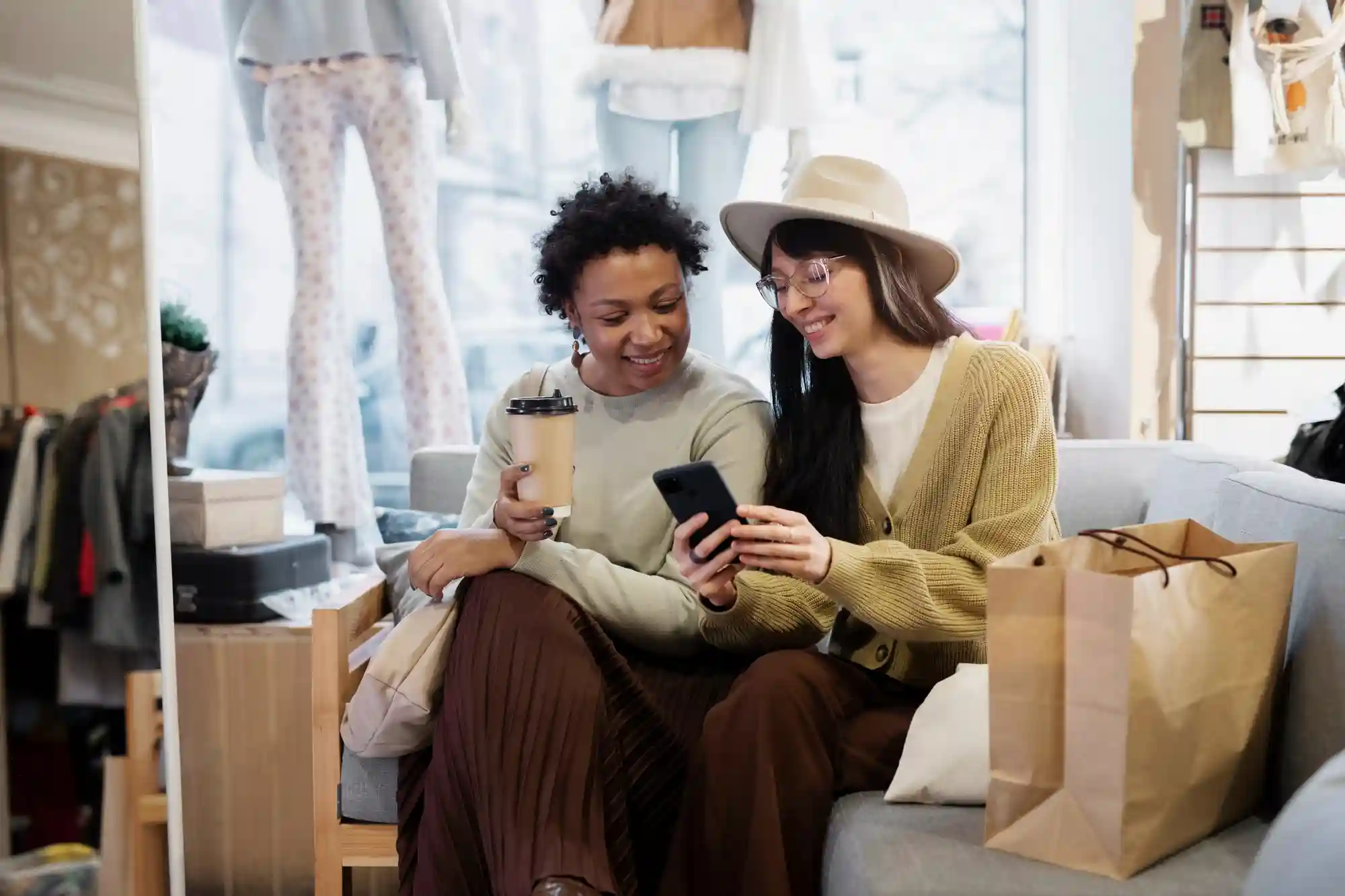 Two friends sit together in a boutique, smiling while using a smartphone to browse digital coupons and deals.