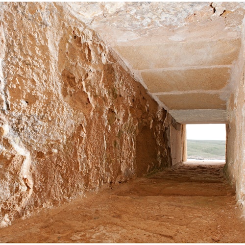 Utsikt över en grovhuggen stenkorridor som leder till solljus, med jordgolv och texturerade stenväggar. Ljuset i slutet visar ett avlägset landskap.