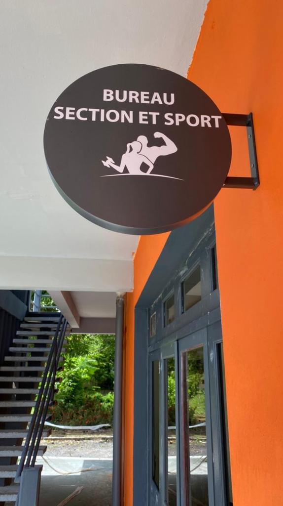 Signalétique Exterieur « Bureau Section et Sport » avec une silhouette en train de faire des flexions et un haltère sur un mur orange vif, à côté de fenêtres et d'escaliers modernes gris foncé.