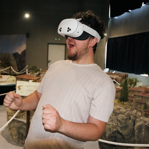 一名佩戴虛擬實境頭戴裝置的人看起來十分興奮，雙手緊握成拳，身處一個陳列各種模型和展品的房間內。