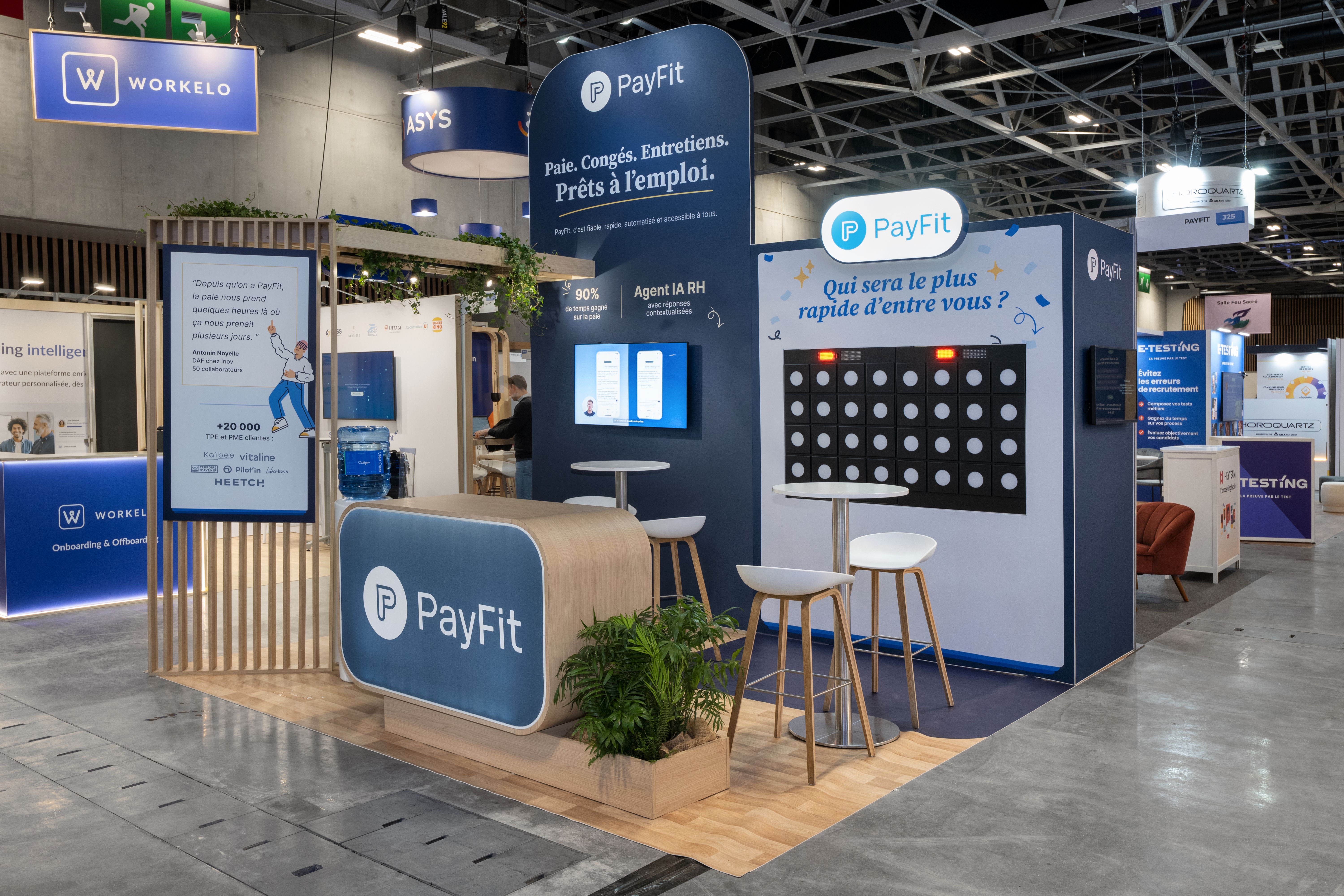 Stand PayFit salon HR Technologies 2026 réalisé par Expace – vue frontale avec arche bois végétalisée, fond bleu marine Agent IA RH, jeu interactif Puissance 4 et comptoir d'accueil arrondi bois et bleu
