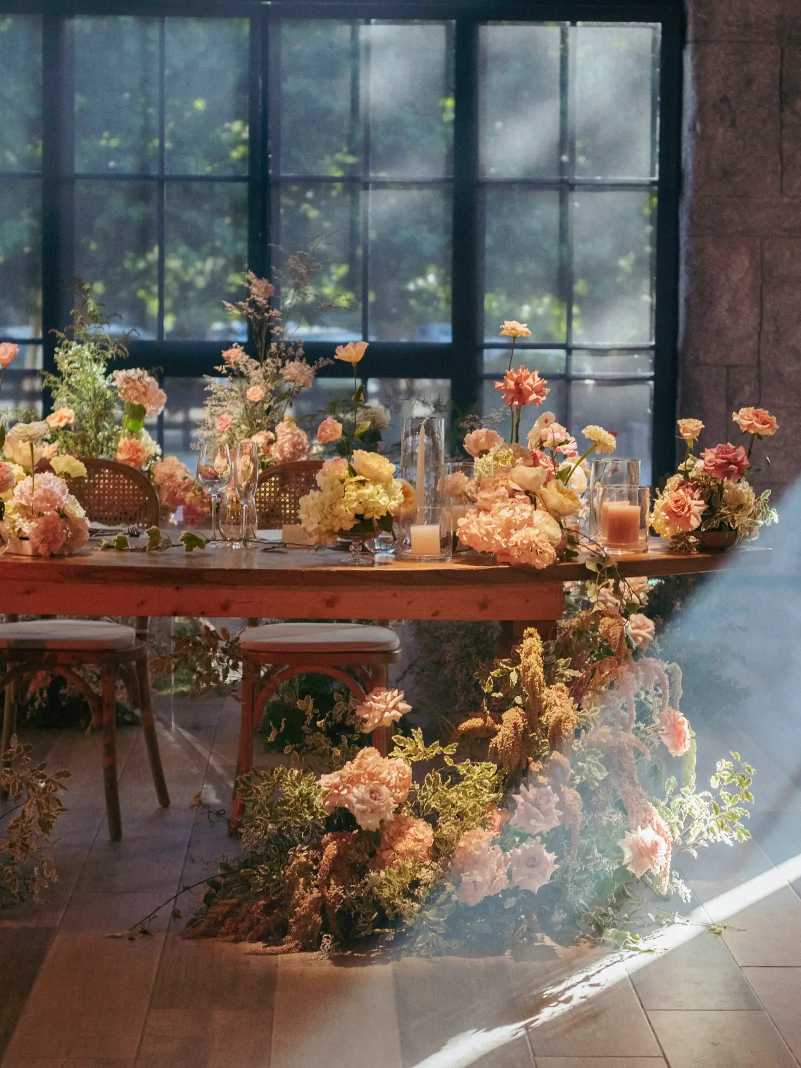 Mesa decorada con flores en tonos durazno y crema, con luz natural filtrándose por las ventanas y arreglos florales que caen hacia el piso.