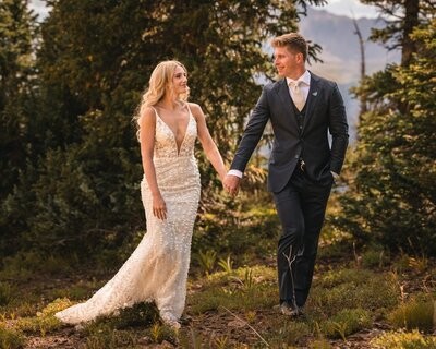 The couple walk hand in hand along a woodland path, smiling at each other.