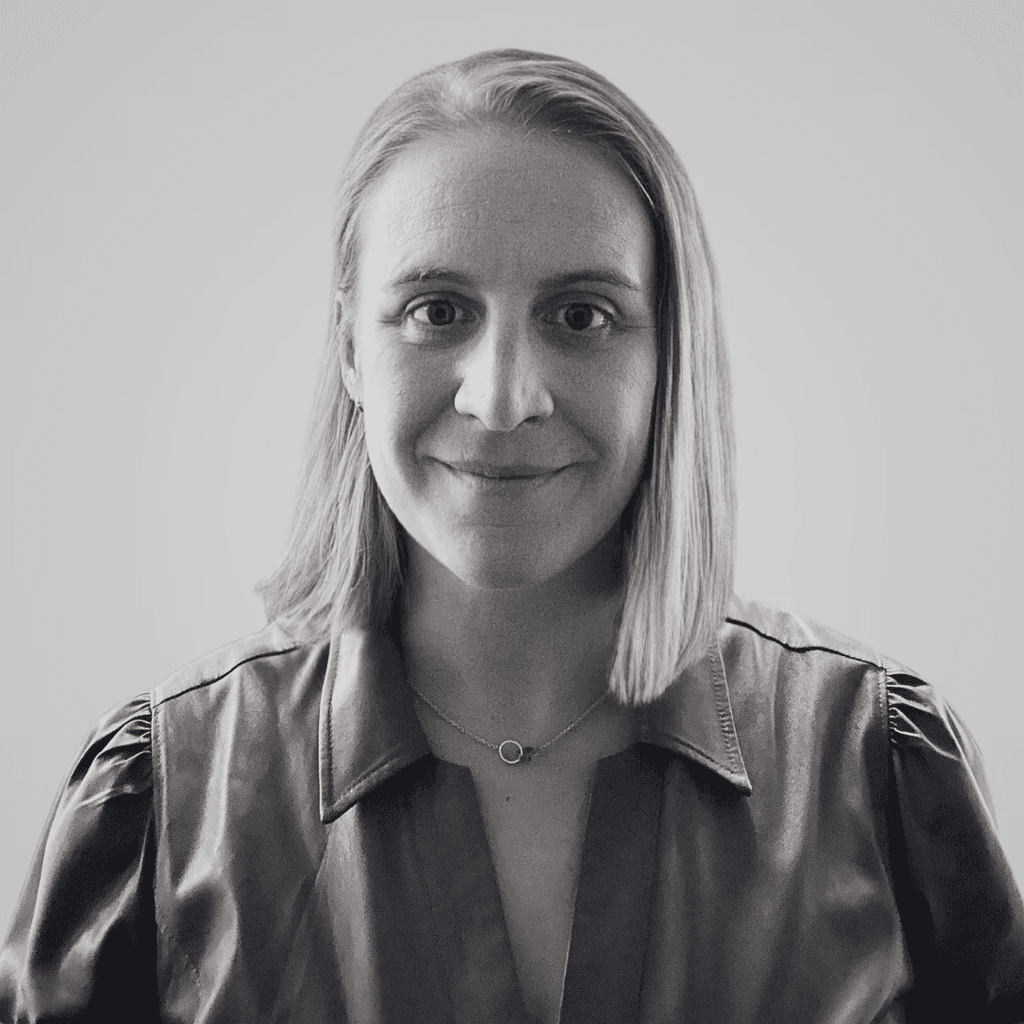 Black and white headshot of a white woman with blonde hair, wearing a dark button-down shirt and a necklace, smiling directly at the camera against a light background. This is the BeyondTrucks Head of Product, Mahriah Alf