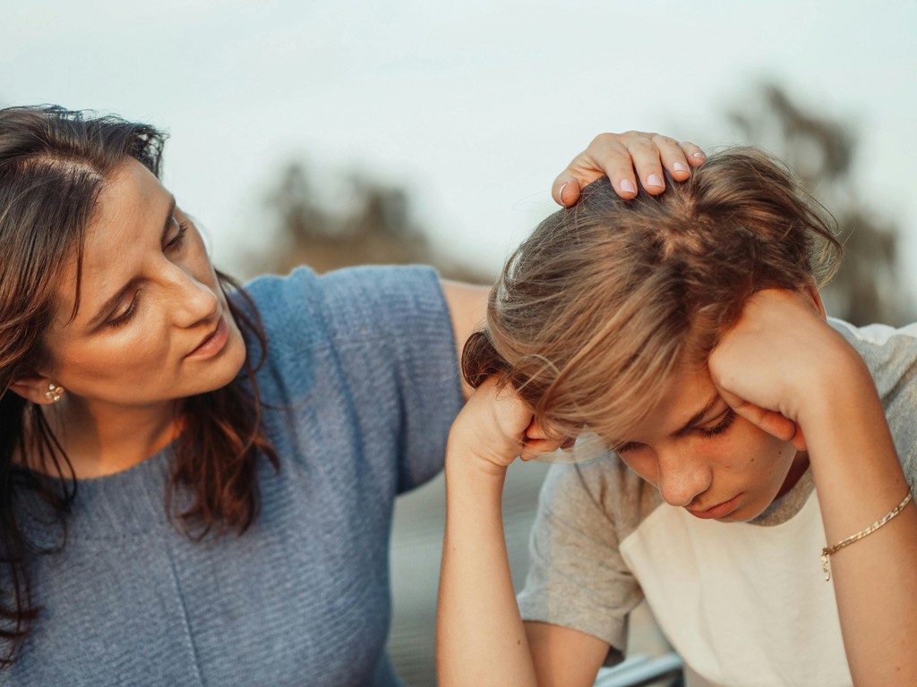 A concerned mother comforts her upset teenage son by gently placing her hand on his head, illustrating a moment of empathy and emotional support during a difficult time.