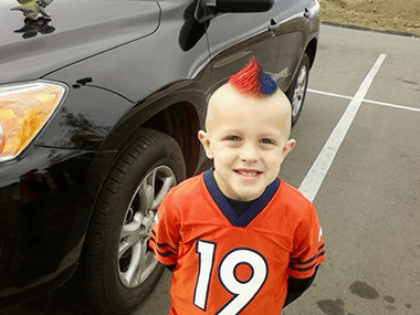 A young Cooper looks up at the camera in a parking lot, he is wearing a Broncos jersey and has a split-dye blue and orange mohawk.