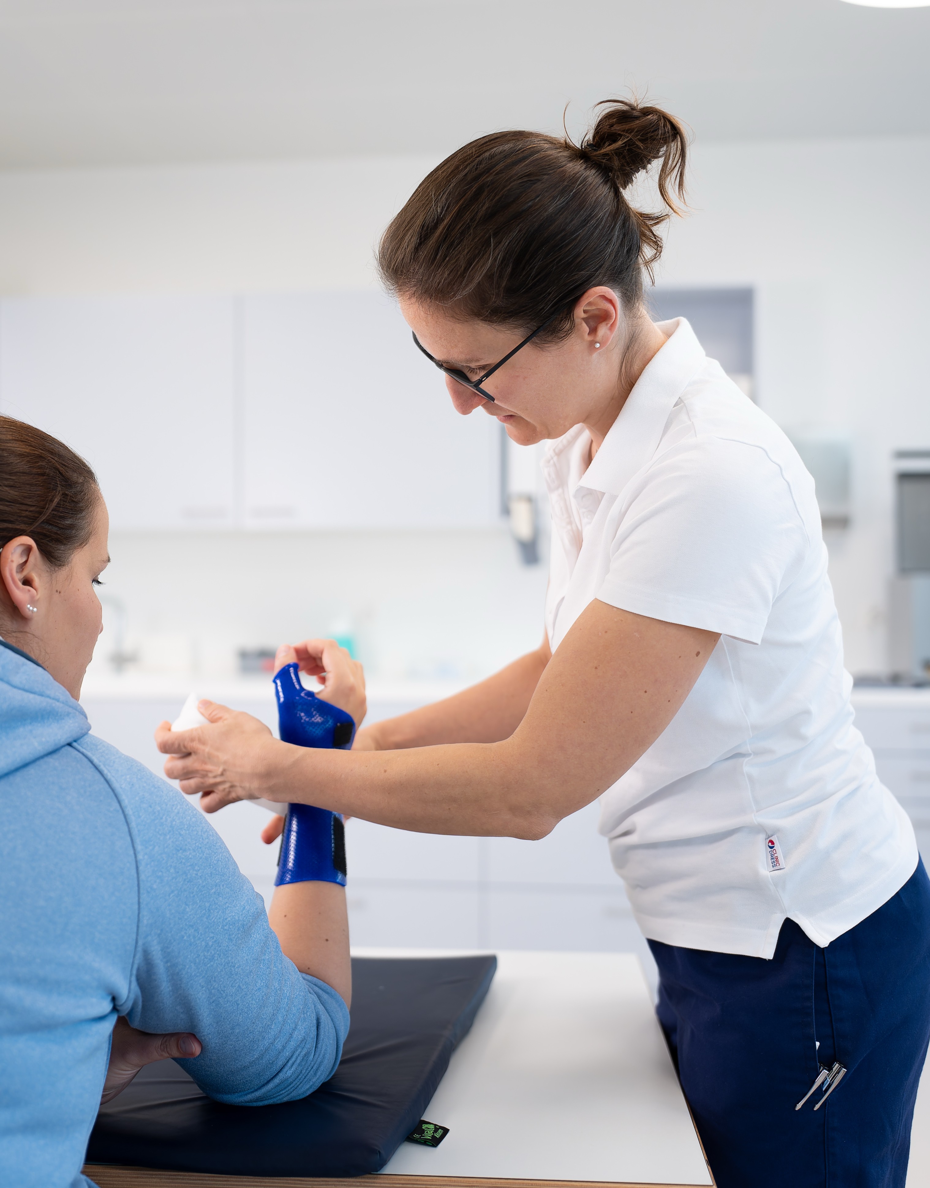 Eine Handtherapeutin im Handzentrum Pratteln legt in einer hellen, klinischen Umgebung einen blauen Verband am Arm einer Patientin an. Die Patientin sitzt in einem blauen Kapuzenpulli, während die Fachperson ein weisses Polo und eine marineblaue Hose trägt.