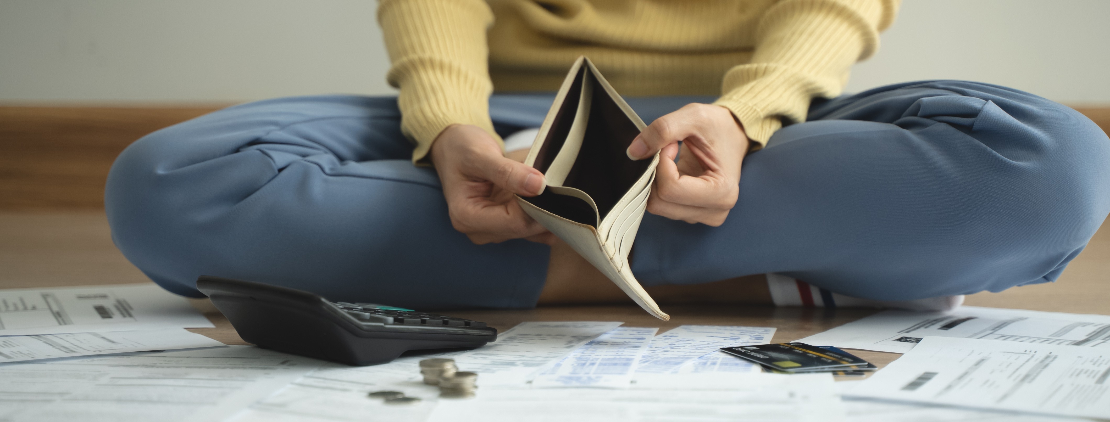 Person sitting on the floor holding an empty wallet, surrounded by bills, a calculator, coins and credit cards.