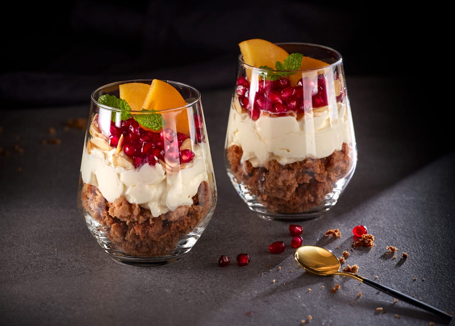 Two elegant glass cups filled with layered dessert showcase a rich combination of crumbled cake, creamy layers, pomegranate seeds, and garnished with peach slices and mint leaves, set against a dark background with a gold spoon adjacent, embodying a delightful Abarel Studio Photo.