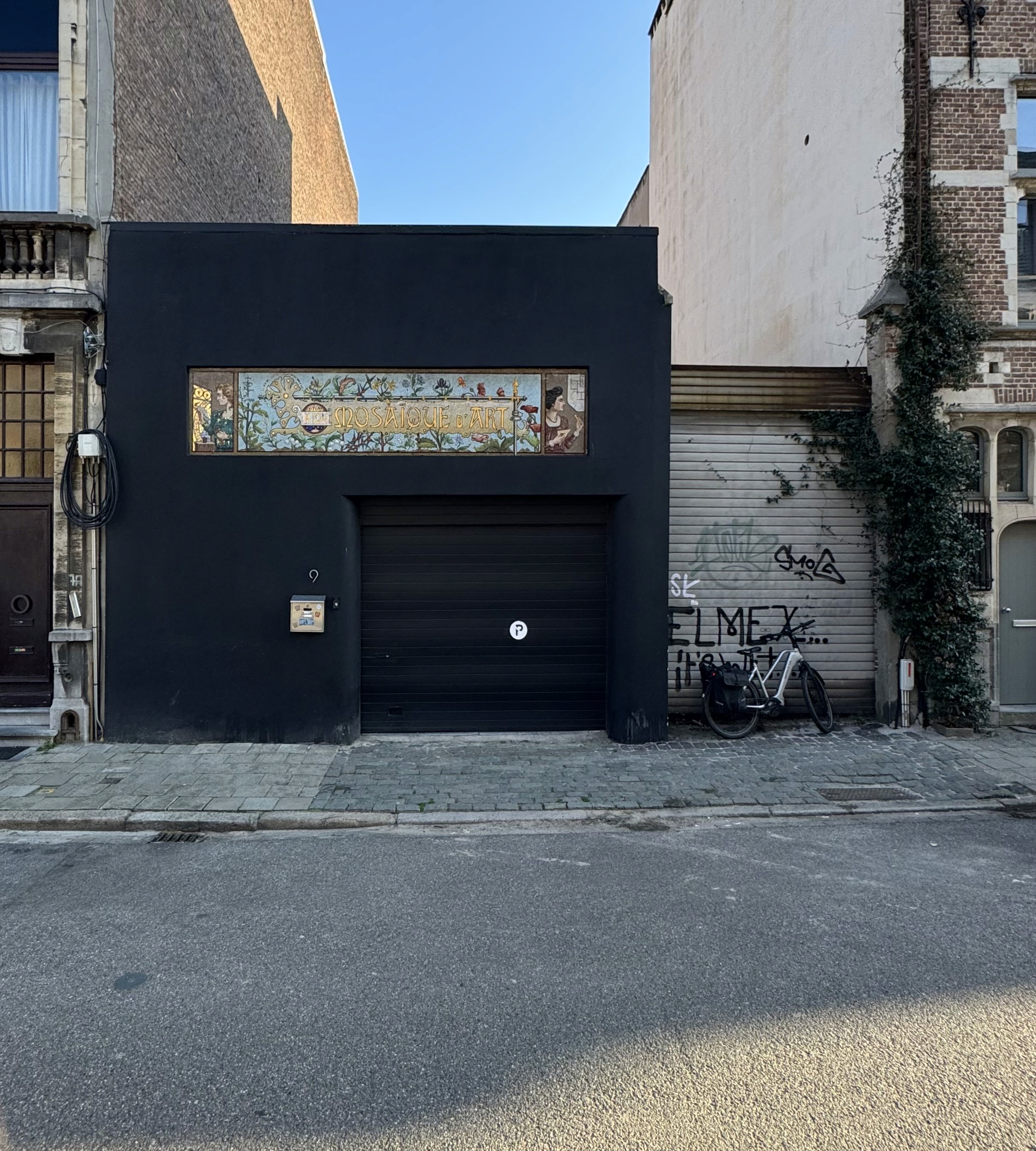 A black garage front with a tiled mosaic sign above the door, on a street in Antwerp