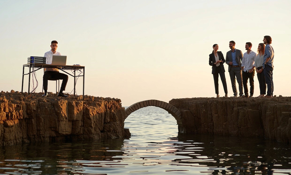 photo of two cliff edges facing each other above calm water at sunrise. On the left cliff, an IT professional sits at a desk with a laptop and server equipment behind them, focused but approachable.