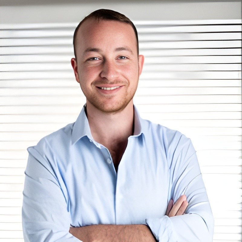 A smiling man with curly hair is sitting at a desk, looking at the camera, with a laptop and a lamp in the background.
