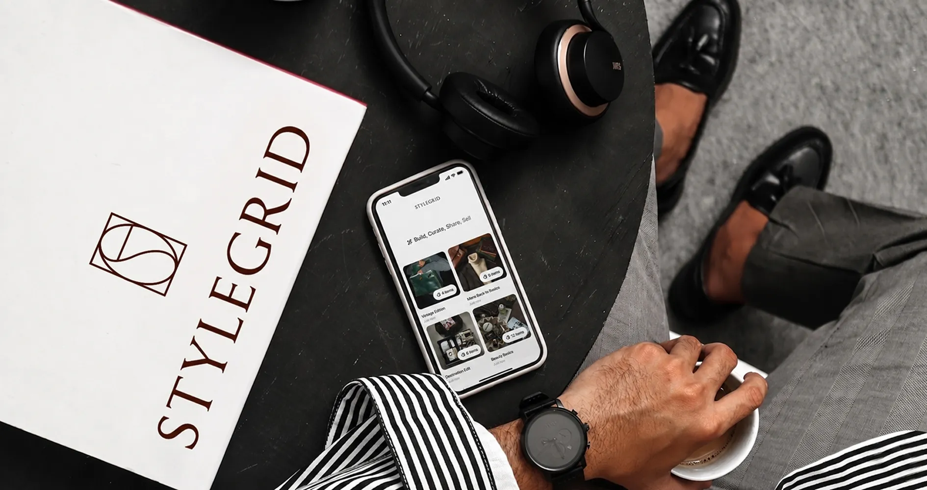 iPhone displaying the StyleGrid app's curated edits on a side table, beside a STYLEGRID lookbook, headphones and coffee.