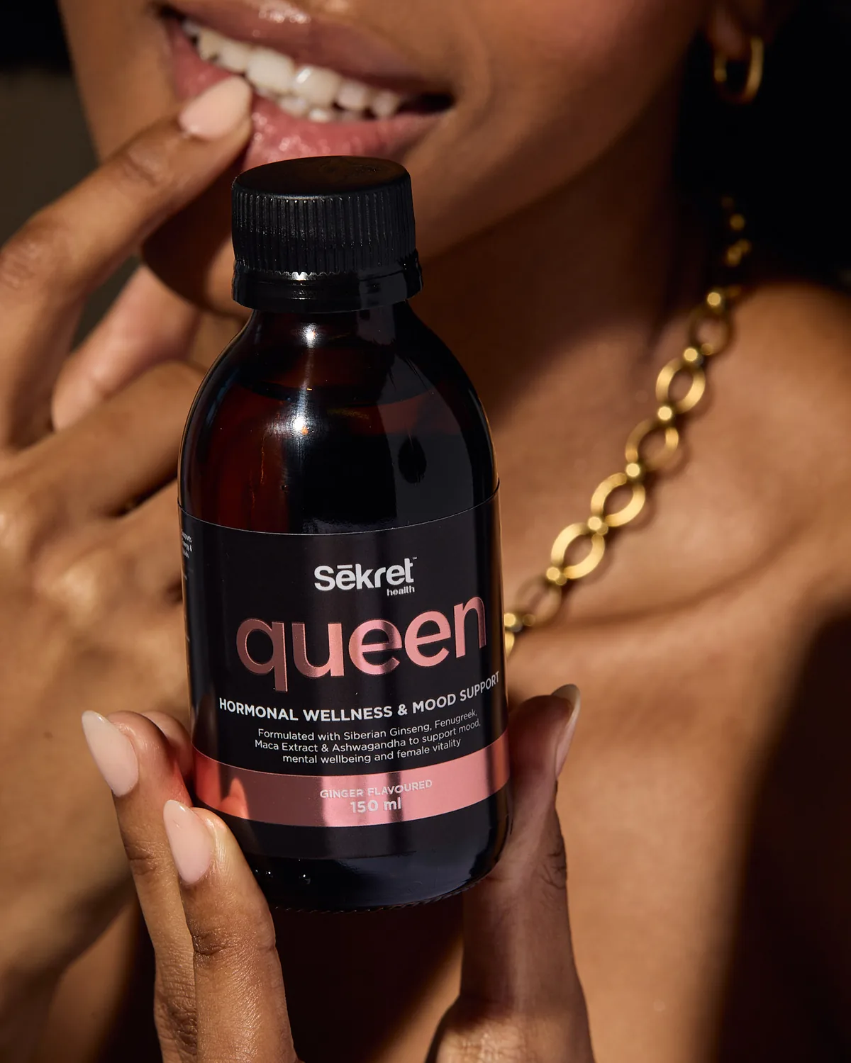 A woman in a gold chain smiles behind a bottle of Sēkret Queen syrup