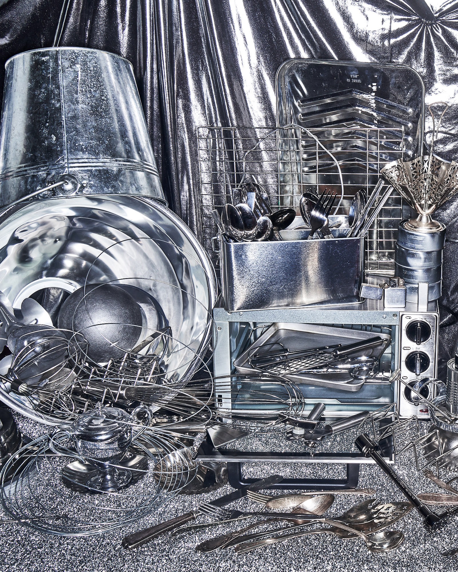 Vertical silver still life assembled from reflective cookware, utensils, tools, and hardware