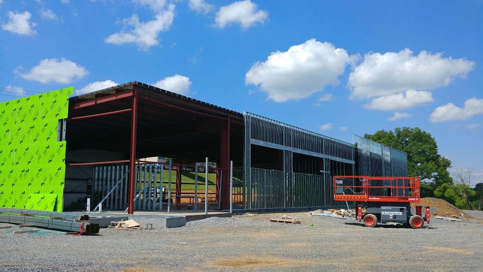 A large building under construction with a green wall, surrounded by gravel and a blue sky with clouds.