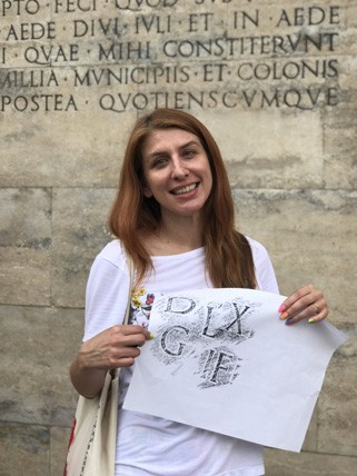 A photo of a girl holding a piece of paper with letters copied from a stone carved wall behind her.