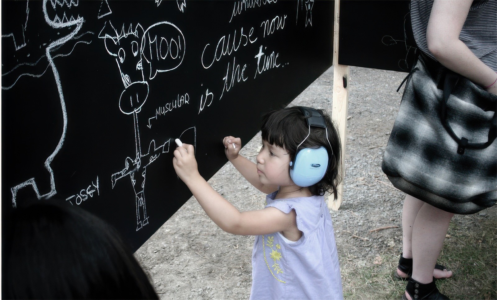 A small child in blue ear protectors draws in chalk on a black board already filled with hand-drawn figures and words.