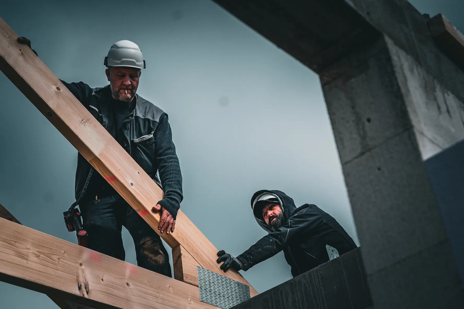 Zwei Bauarbeiter in Schutzkleidung montieren vor einem bewölkten Himmel Holzbalken an einer Gebäudekonstruktion.