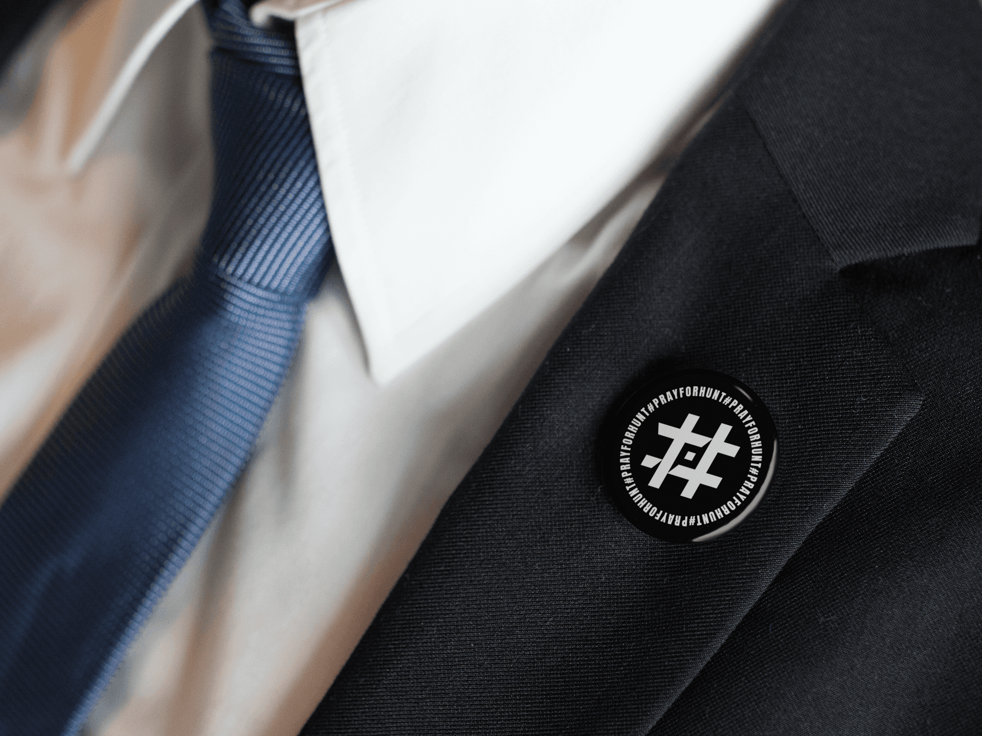 A close-up of a person wearing a black suit with a white dress shirt and a dark blue tie. Attached to the lapel is a round black button featuring the '#PrayForHunt' logo. The button has a white hashtag symbol with a Shuriken element in the center, surrounded by the text '#PrayForHunt' repeated in a circular pattern. The design contrasts sharply with the suit, making the button stand out as a strong statement piece.