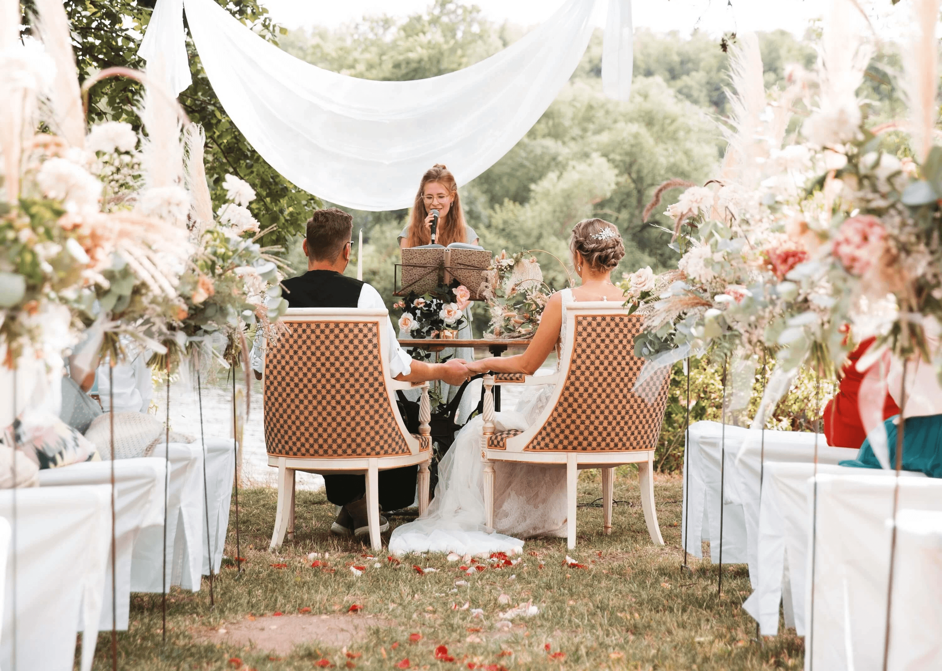 Brautpaar bei Hochzeitszeremonie unter goldenem Bogen, umgeben von Blumen. Frau liest, Braut trägt Schleier, Bräutigam im Anzug.