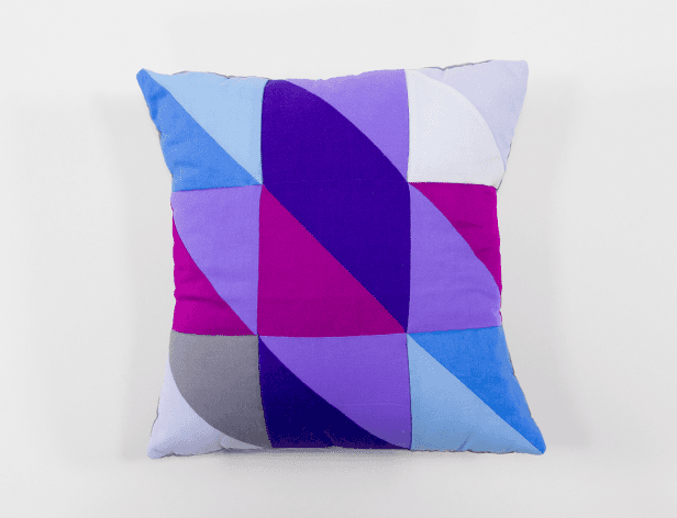 A square throw pillow with a geometric pattern composed of overlapping triangles in shades of blue, purple, pink, and white. The pillow rests on a white surface against a plain white background.
