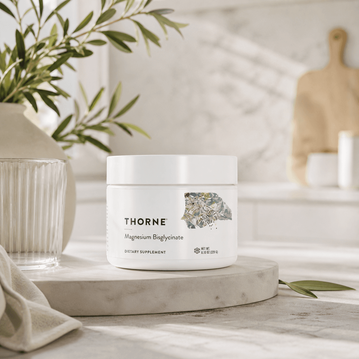 A white jar of Thorne Magnesium Bisglycinate supplement sits on a marble surface. Soft light, a leafy plant, and kitchen decor create a serene, natural ambiance.