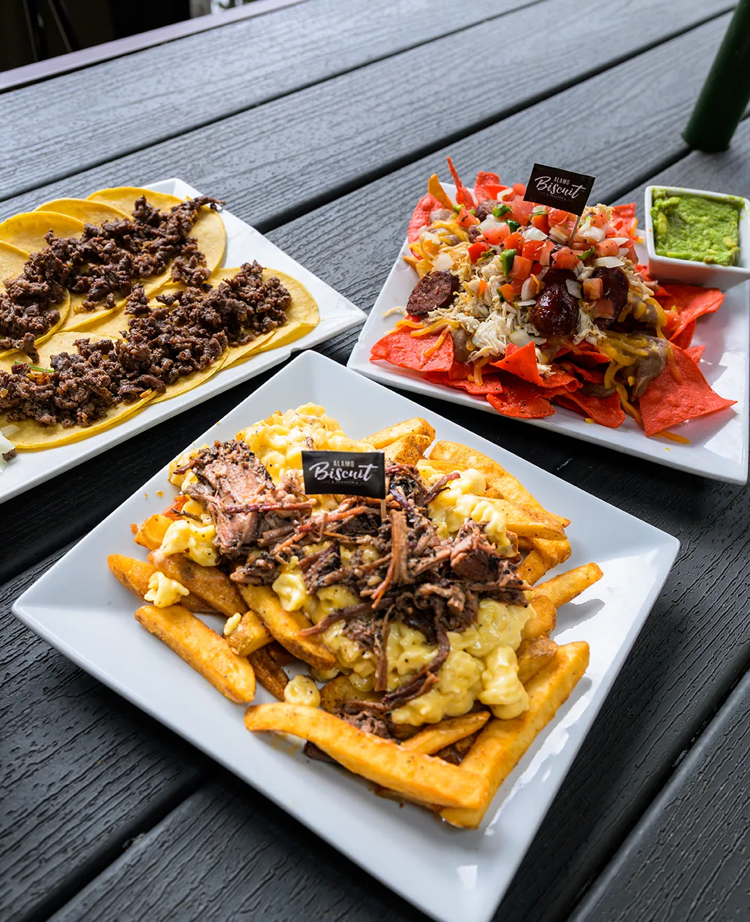 Three shareable plates from Alamo Biscuit on a dark patio table — pulled pork fries with scrambled egg, brisket nachos with guacamole, and beef tostadas