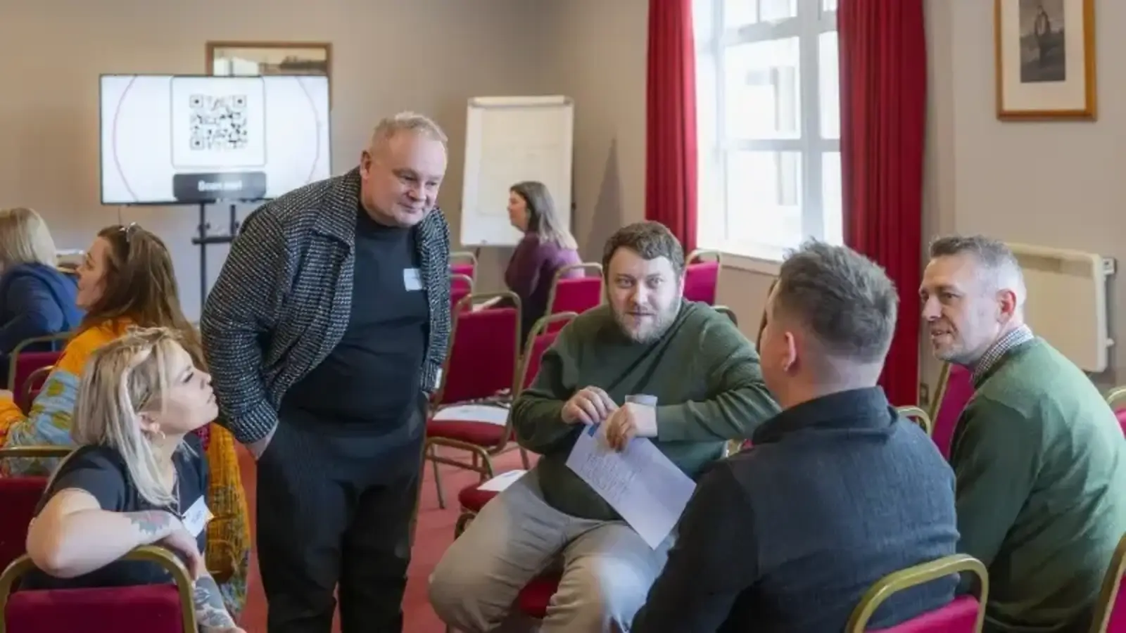 A group of people engaged in a lively discussion in a meeting room with red chairs. A screen displays a QR code in the background, and the mood is collaborative.