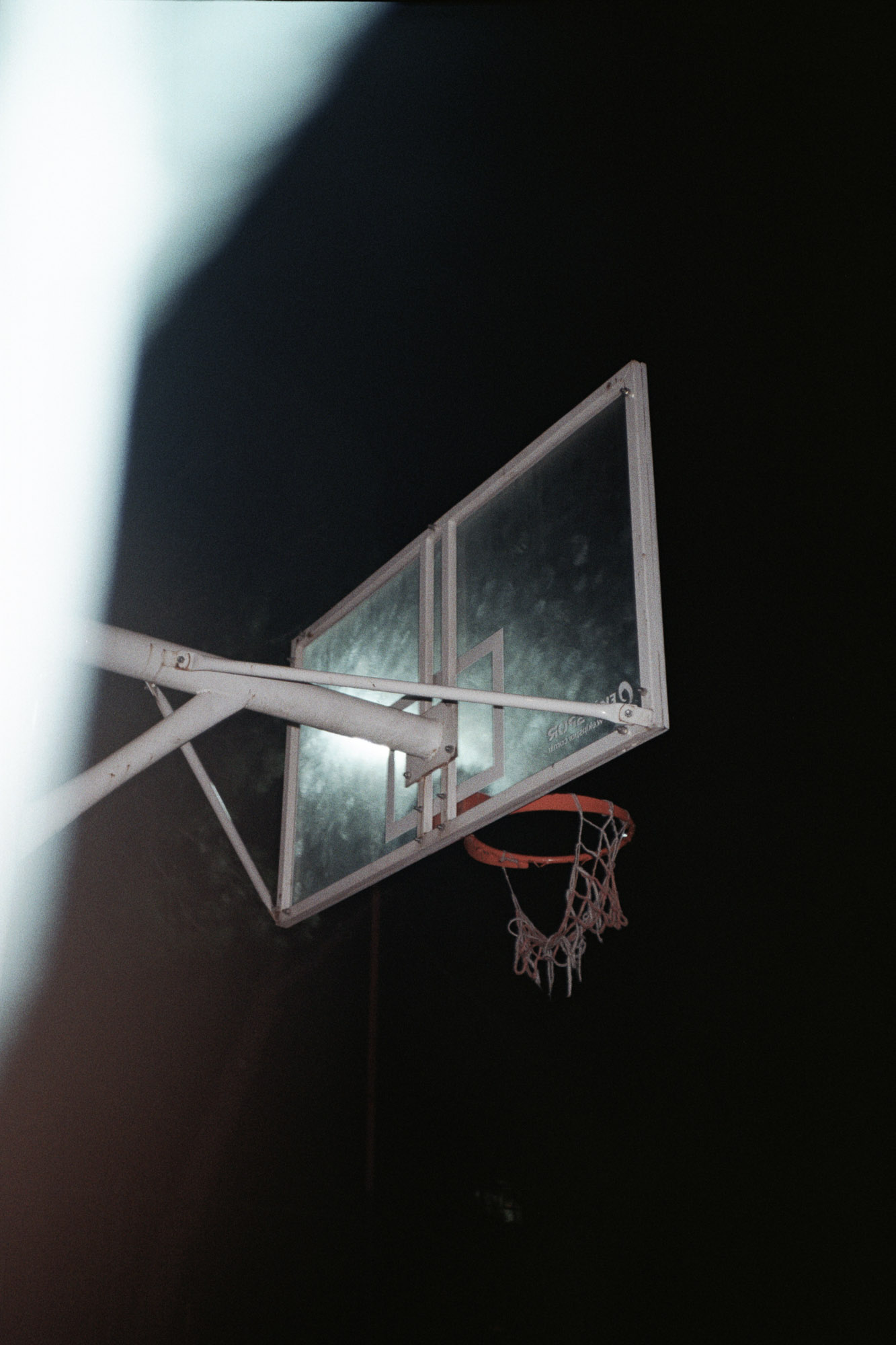 A dimly lit basketball hoop with a tattered net stands isolated against a dark night sky, illuminated by a single overhead light.