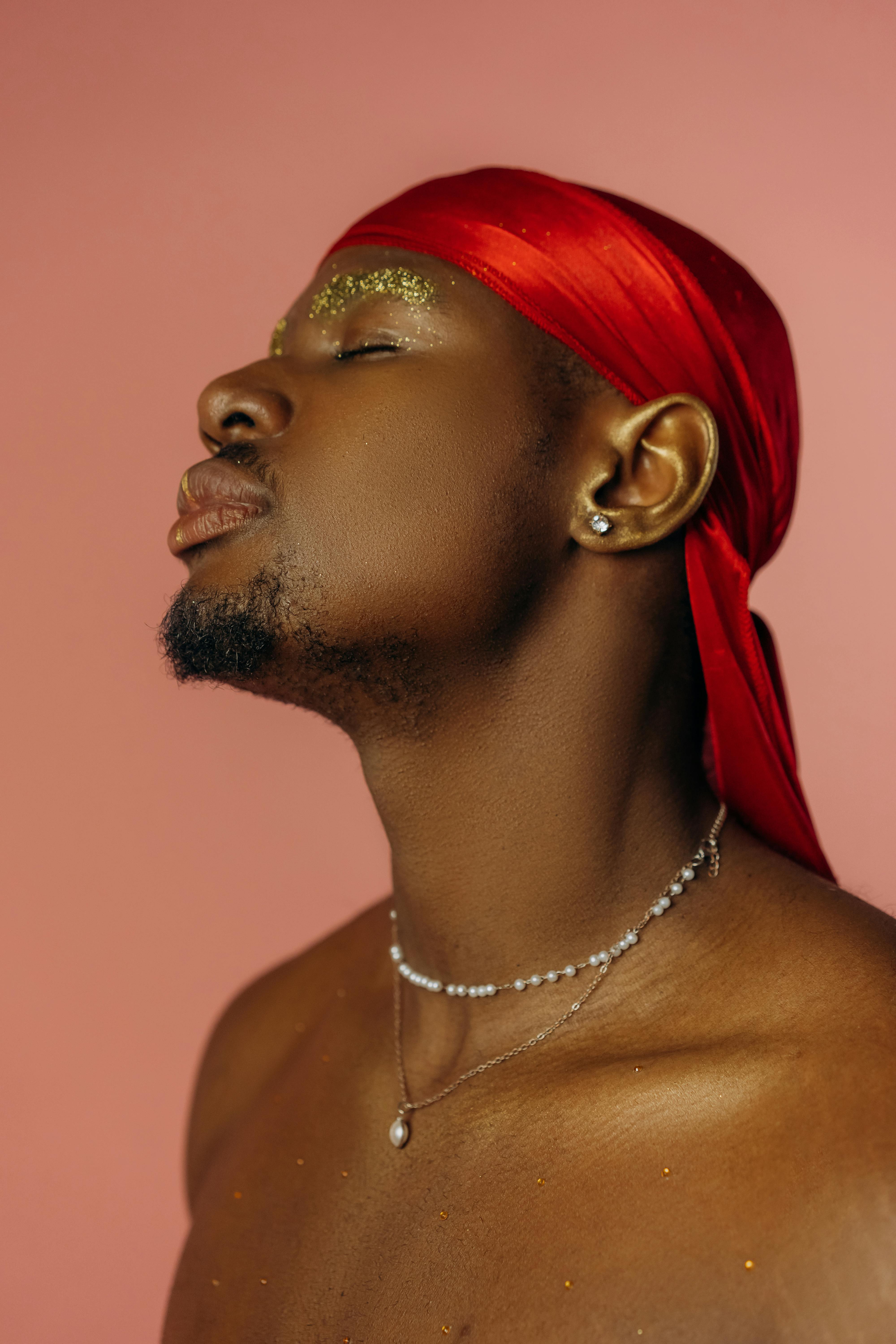 Man in a red durag and necklaces with glitter on his eyelids and chest.