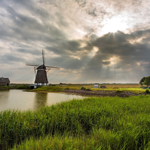 A windmill stands near a pond surrounded by green grass under a cloudy sky with sunlight breaking through.