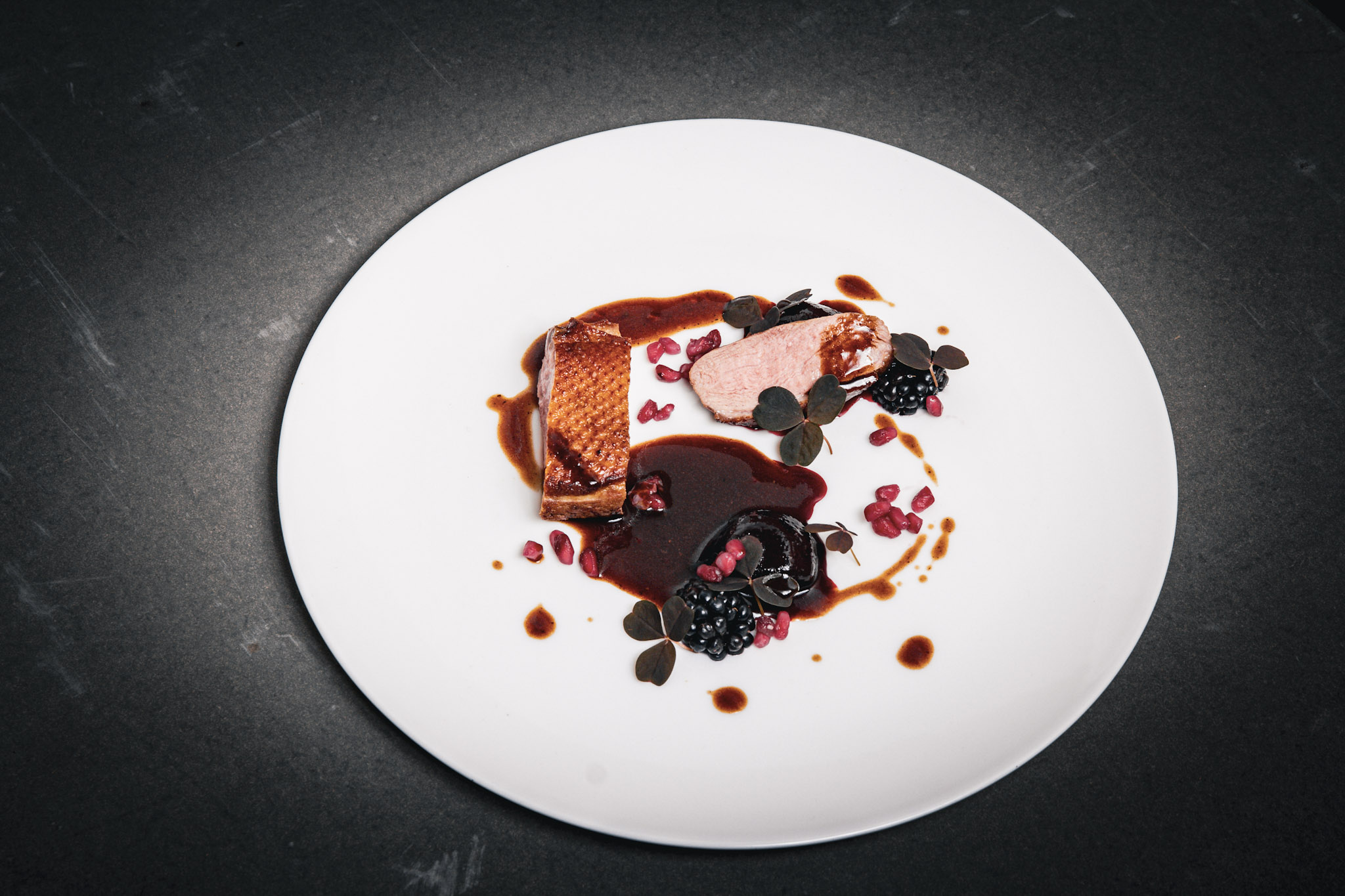 Top-down view of sliced duck breast with crisp skin placed over a pool of dark blackberry sauce, accompanied by glazed beetroot, fresh blackberries, and scattered pomegranate seeds on a white plate.