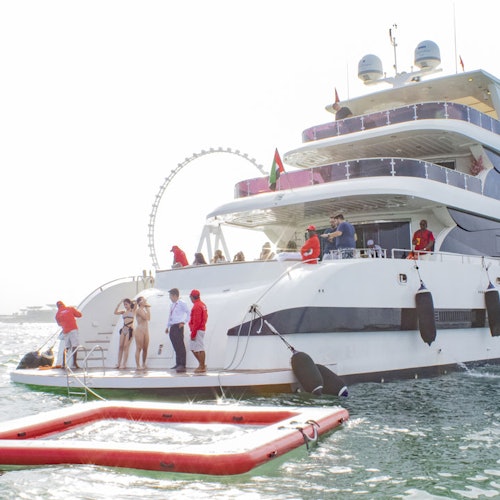 一班人喺一艘大型遊艇上，擁有多個甲板，停靠喺水面上，旁邊有一個紅色浮台，背後有一個摩天輪。