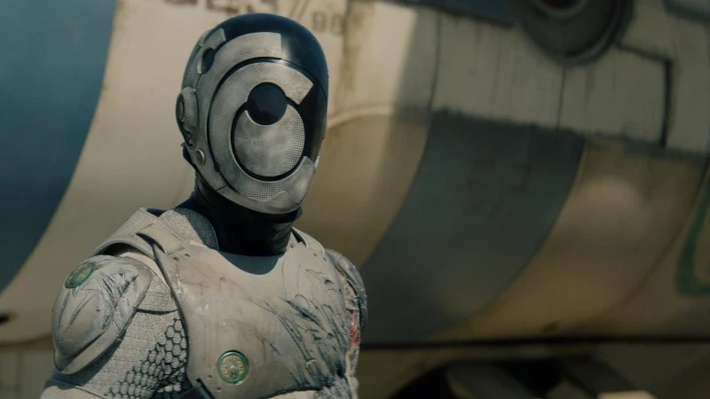 A futuristic humanoid robot with a metallic suit stands in front of an aircraft. The robot's face is featureless, displaying a large circular design. The background shows the side of a weathered plane with blue and white stripes.