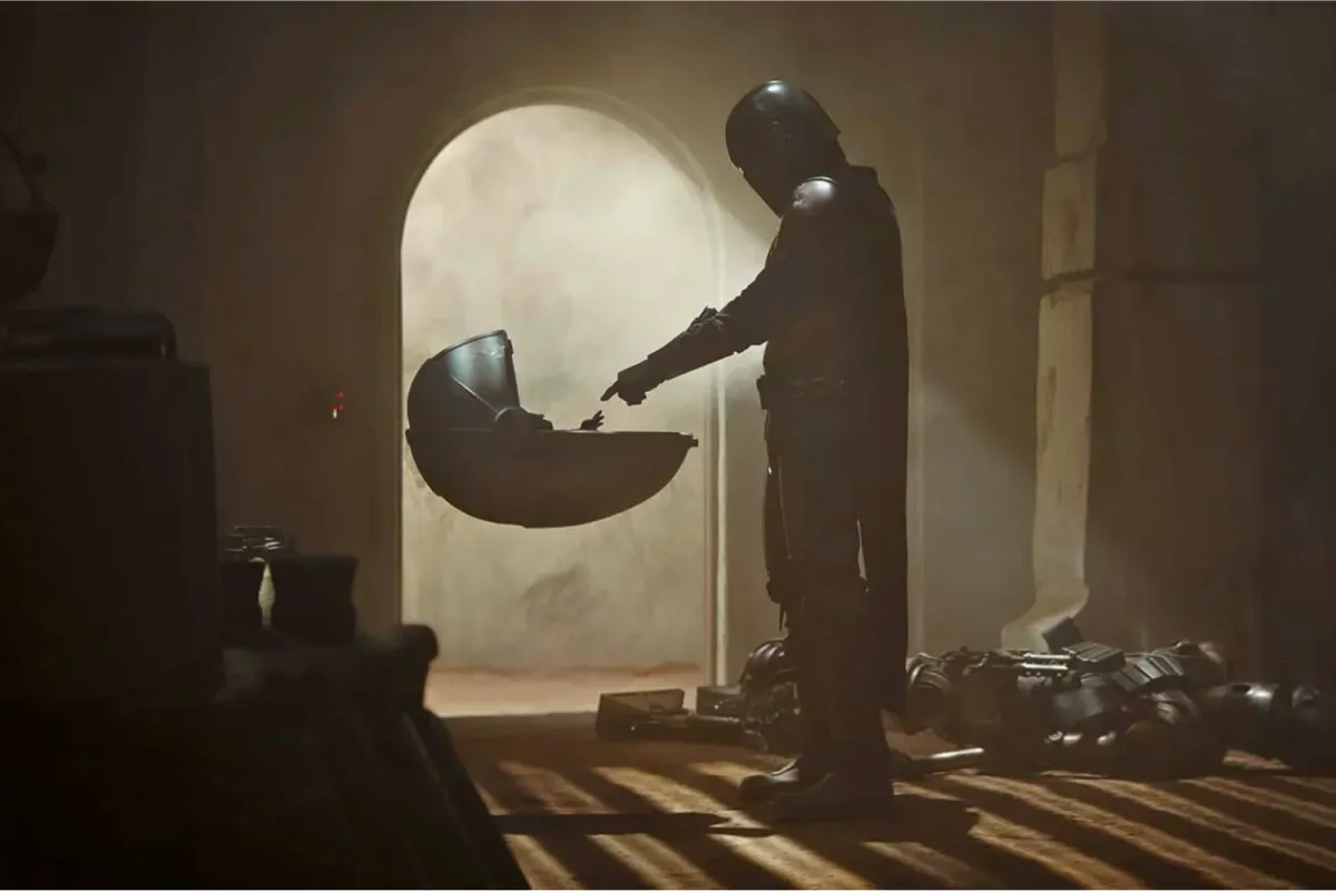 The Mandalorian, clad in full beskar armor, reaches out to Grogu, who sits inside his floating pram. A warm light from an arched doorway silhouettes their touching moment, highlighting the beginning of their bond in The Mandalorian.