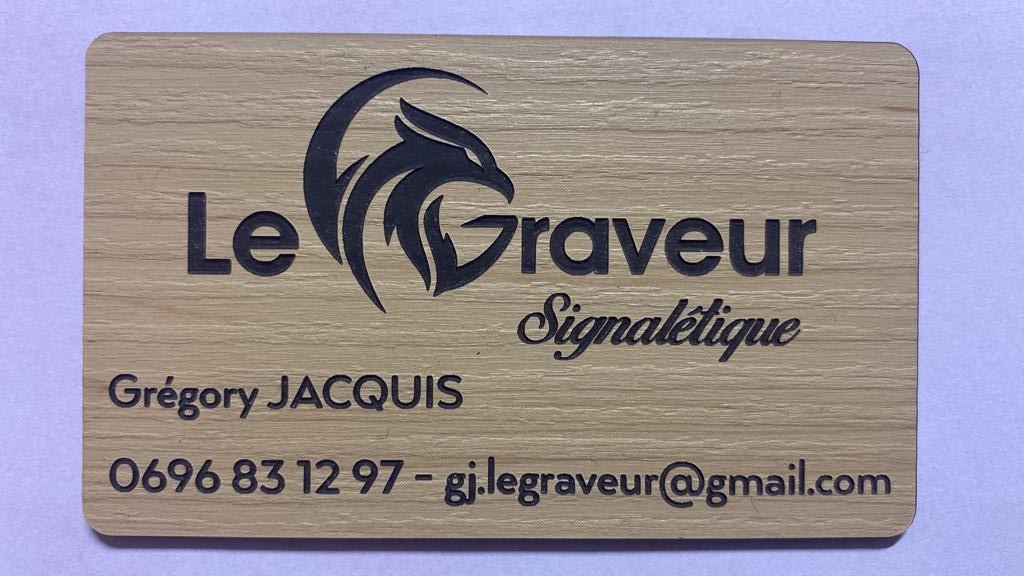 Une carte de visite en bois arbore un logo stylisé représentant un phénix accompagné du texte « Le Graveur Signalétique ». Elle comprend les coordonnées de l'entreprise dans un style professionnel.