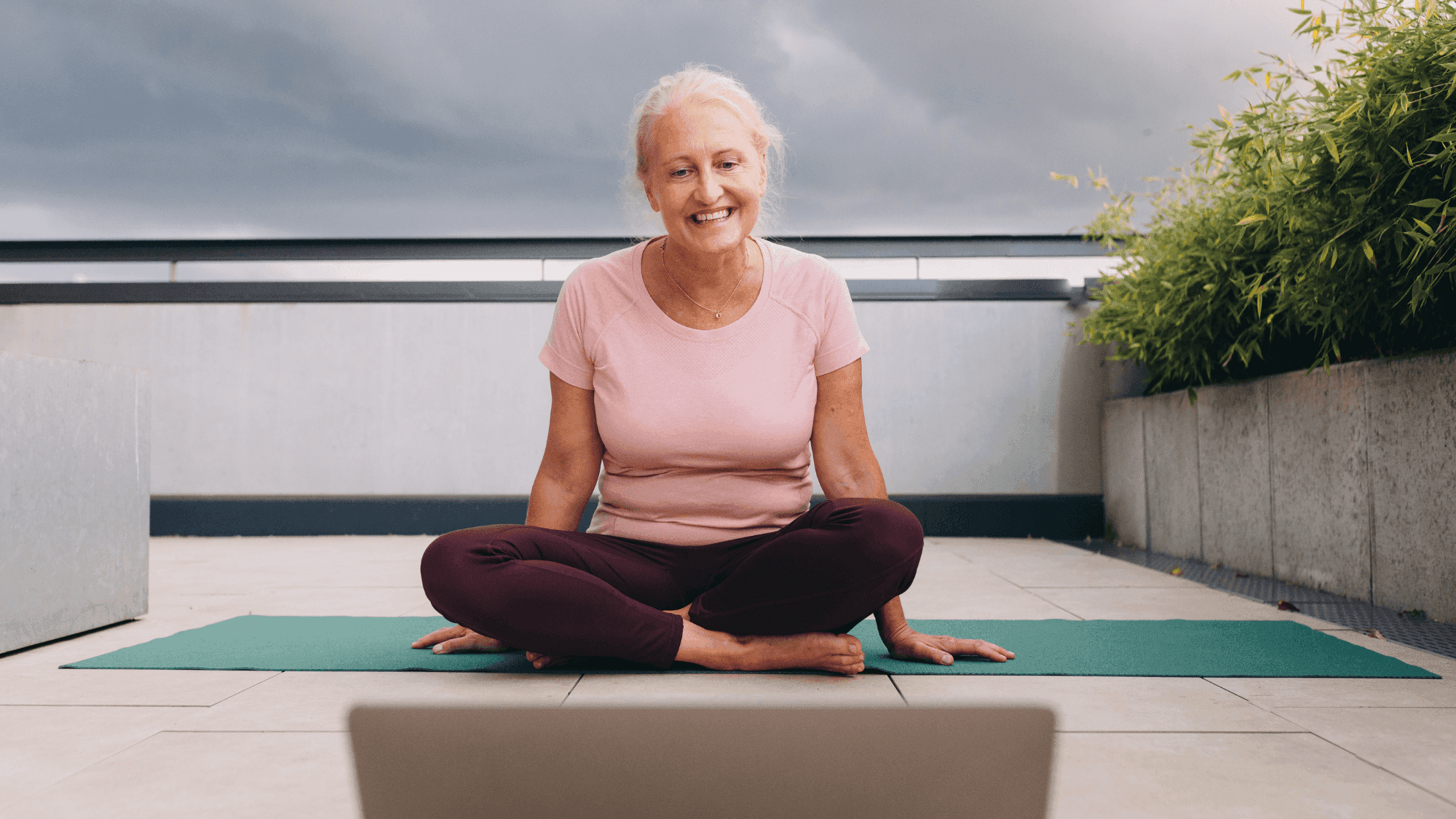 Smiling senior woman in Easy Pose (Sukhasana) on green yoga mat, preparing for online yoga class with laptop, demonstrating accessible yoga practice on outdoor patio