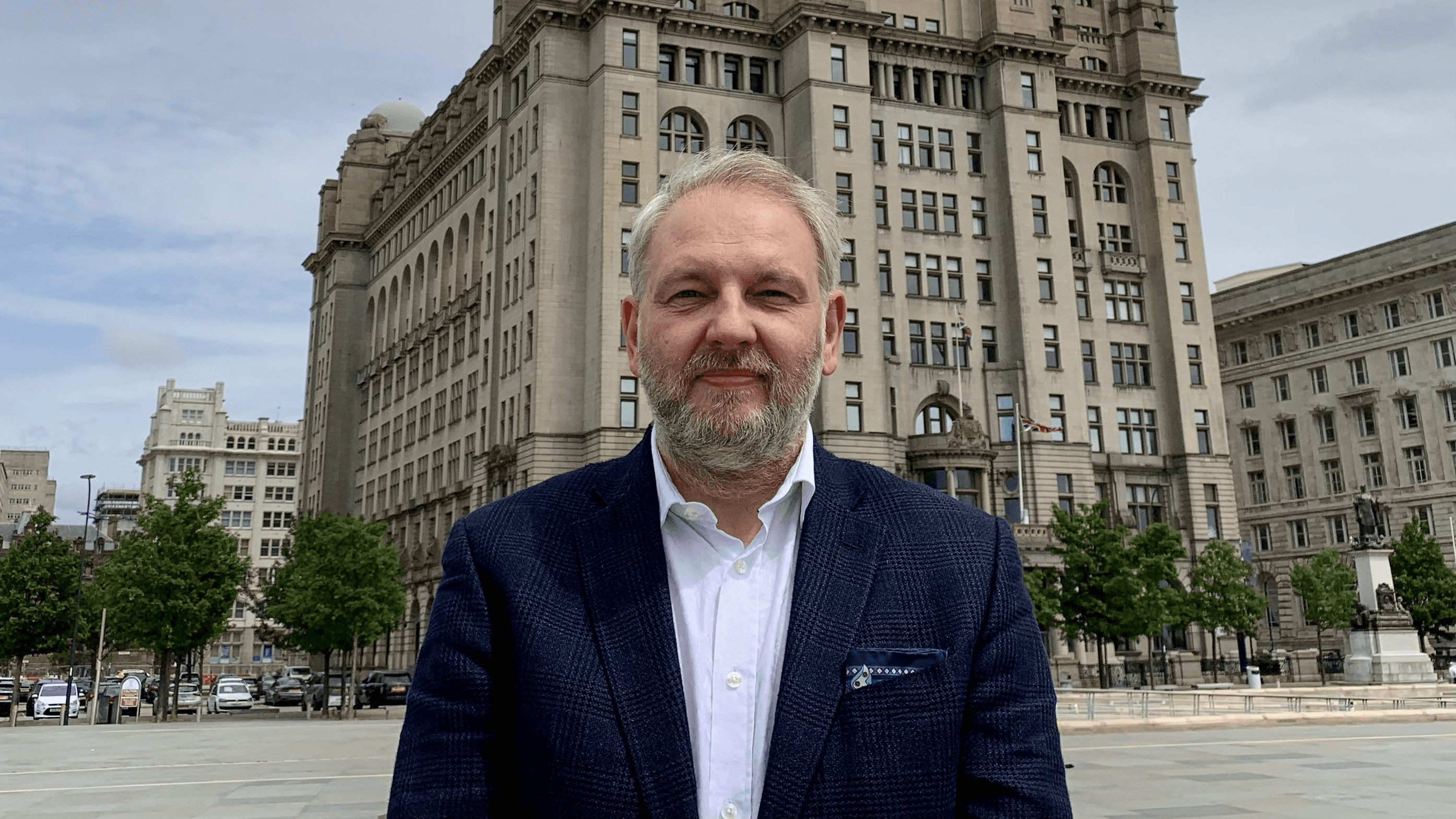A smiling man with short hair stands with arms crossed, wearing a dark shirt against a neutral background.