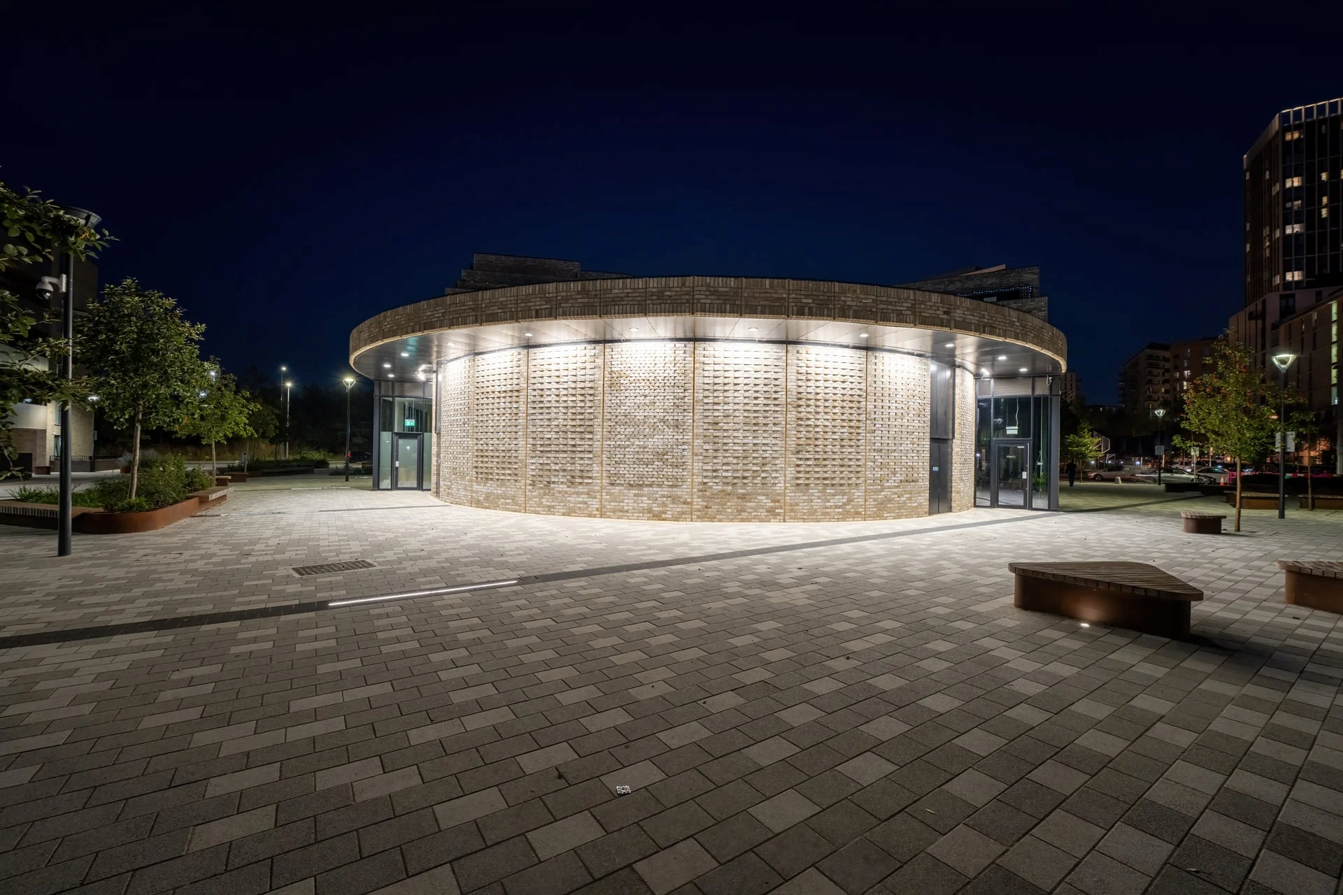 Night-time drone photography capturing the illuminated facade of a modern circular brick building and public plaza