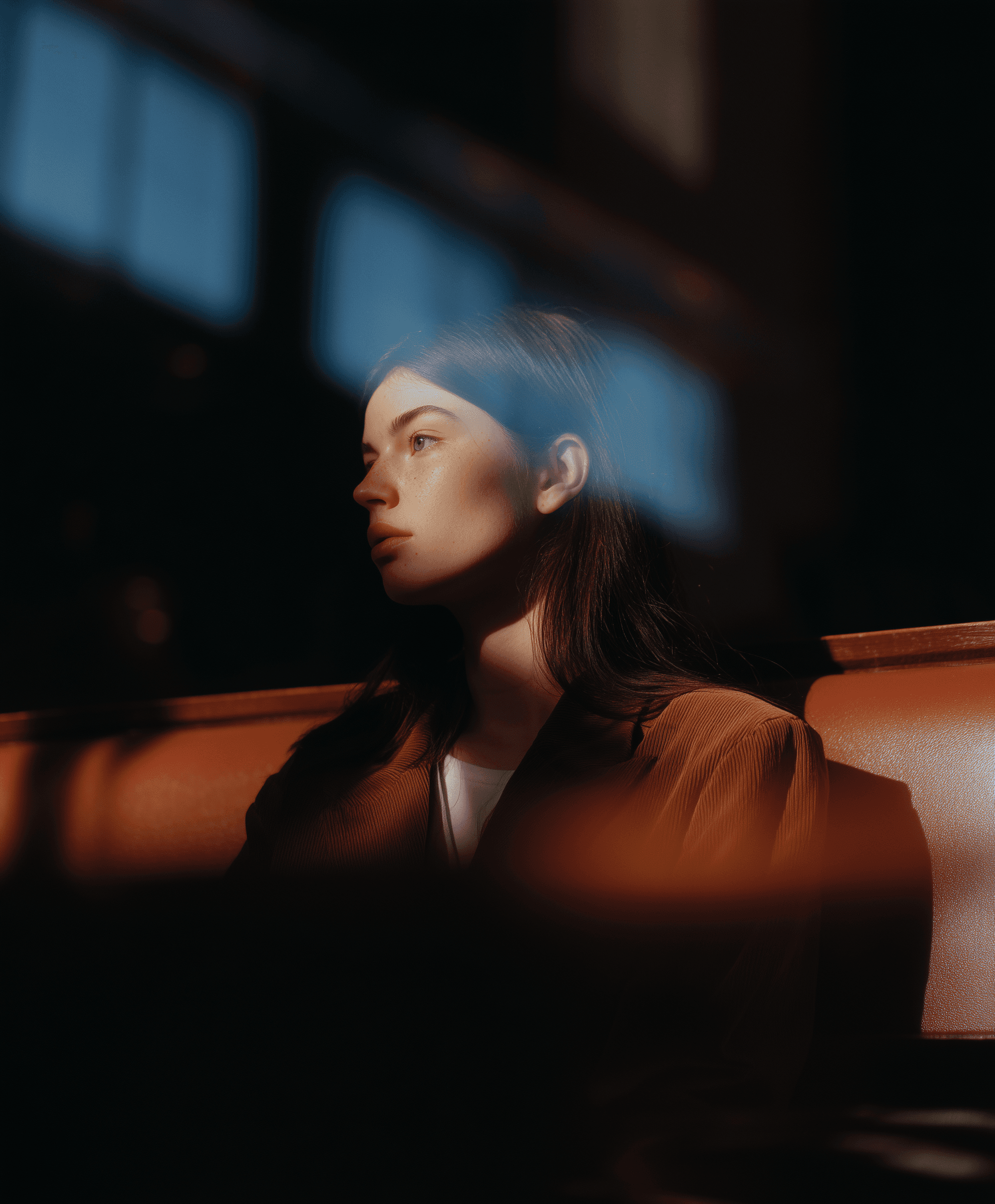A contemplative young woman in warm lighting, wearing a brown jacket, gazes into the distance in a softly blurred interior setting.