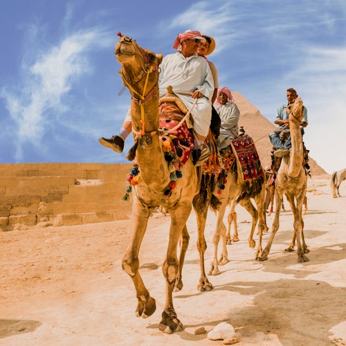 Tourists ride camels, adorned with colorful blankets, near the pyramids under a bright blue sky.