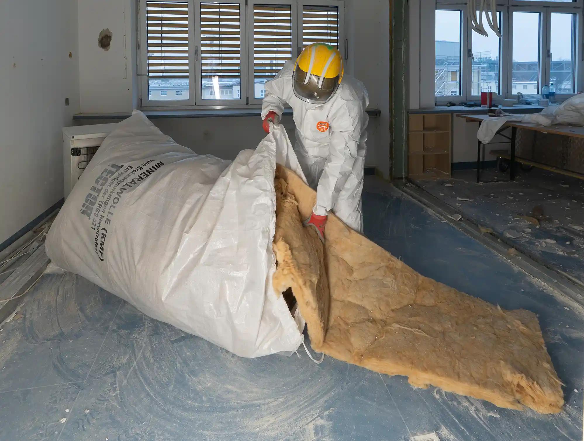 Arbeiter in Schutzanzug bei Schadstoffsanierung beim Verpacken von Mineralwolle in Entsorgungssack im Innenbereich