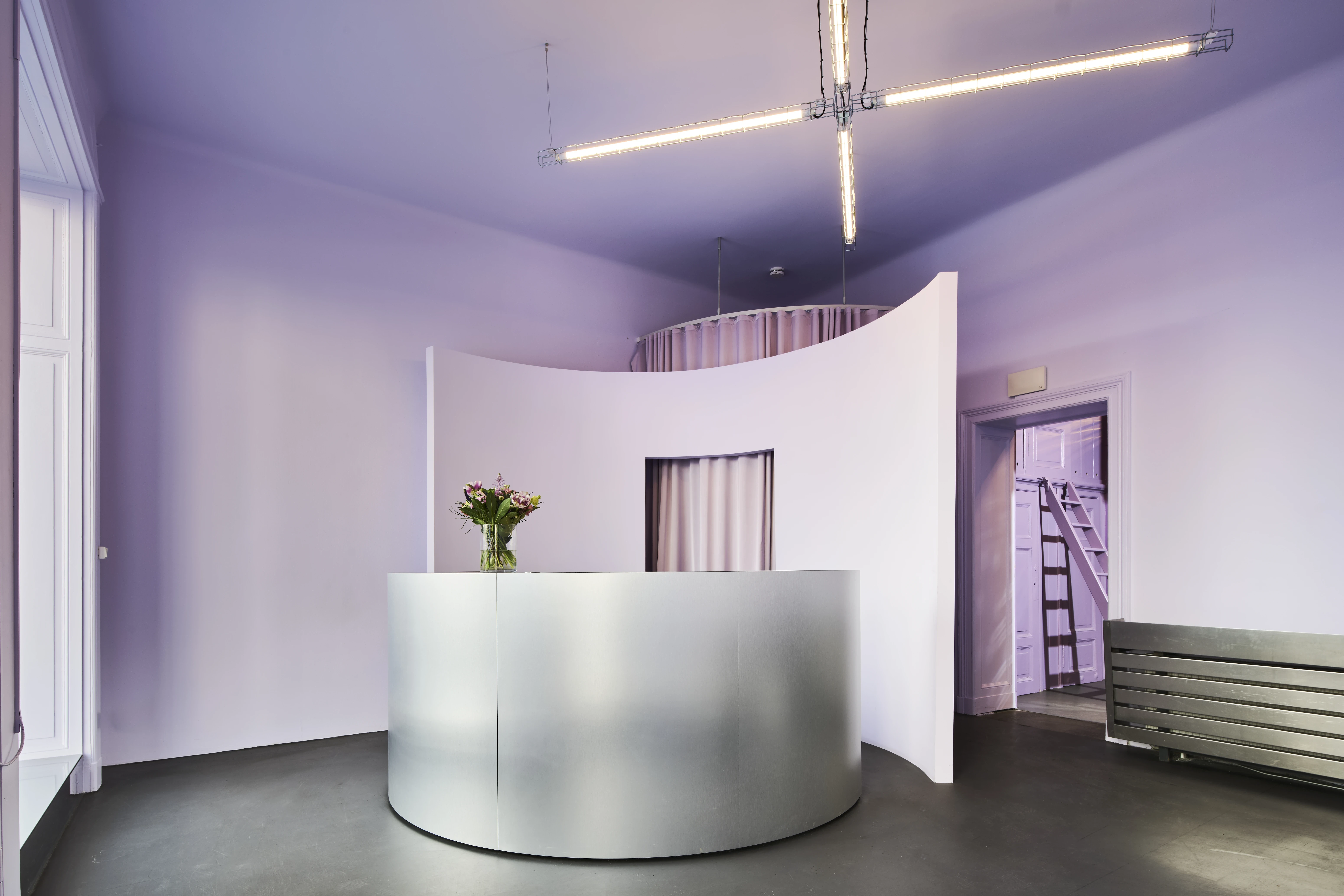 Wide view of the BOLD OFFICE reception area with curved white desk, lavender walls and ceiling, a flower bouquet on the counter, cross-shaped industrial pendant lights, and a metal bench on the right.