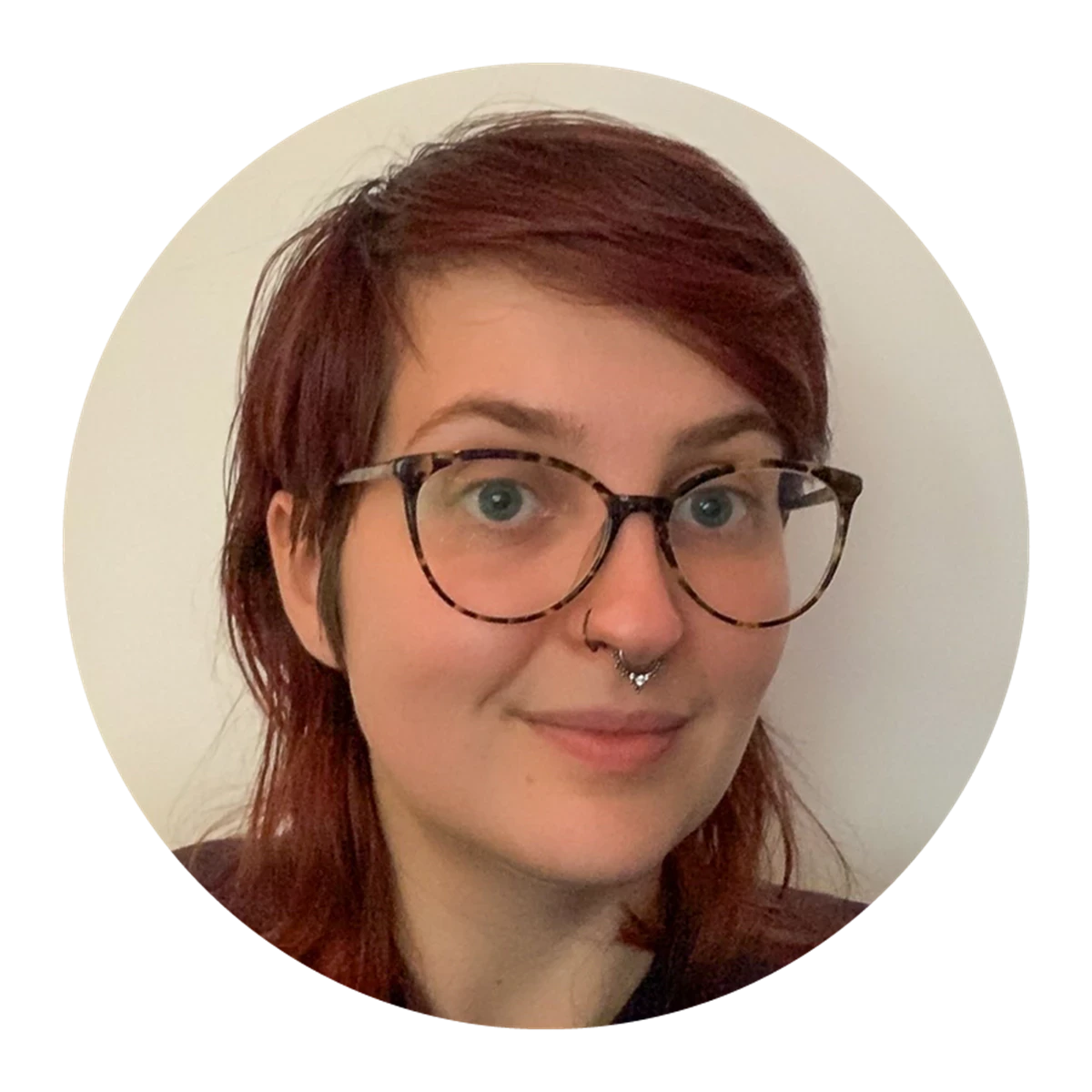 A person with dark red hair and glasses is photographed against a plain light-coloured wall. Part of their glasses frame is visible on one side.