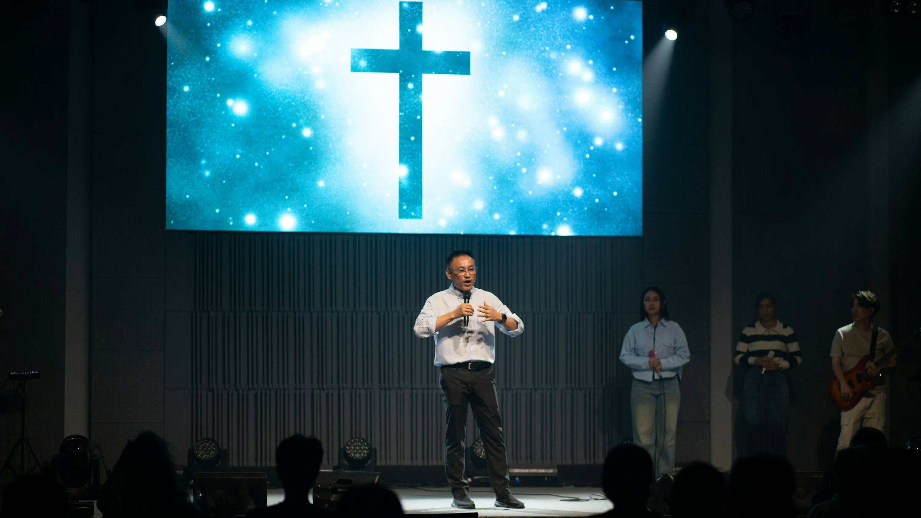 Pastor Daniel Okoro preaching beneath a cross projected on the stage screen, with the band behind him