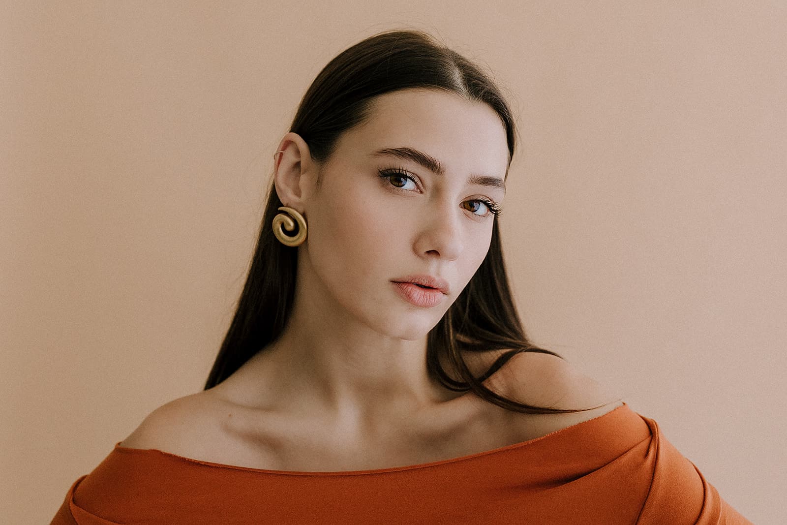 A model with long brown hair wears gold spiral earrings and an off-the-shoulder orange top in a Shreveport natural light studio.